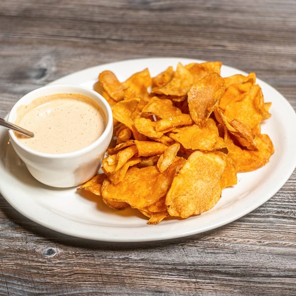 A white plate with a pile of golden-brown potato chips and a small cup of creamy dipping sauce.