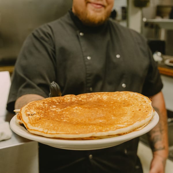 A smiling chef in a black uniform holds out a plate piled high with golden pancakes.