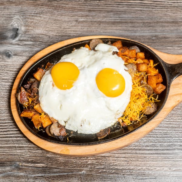 Breakfast skillet with two fried eggs, sausage, potatoes, and shredded cheese on a wooden table.