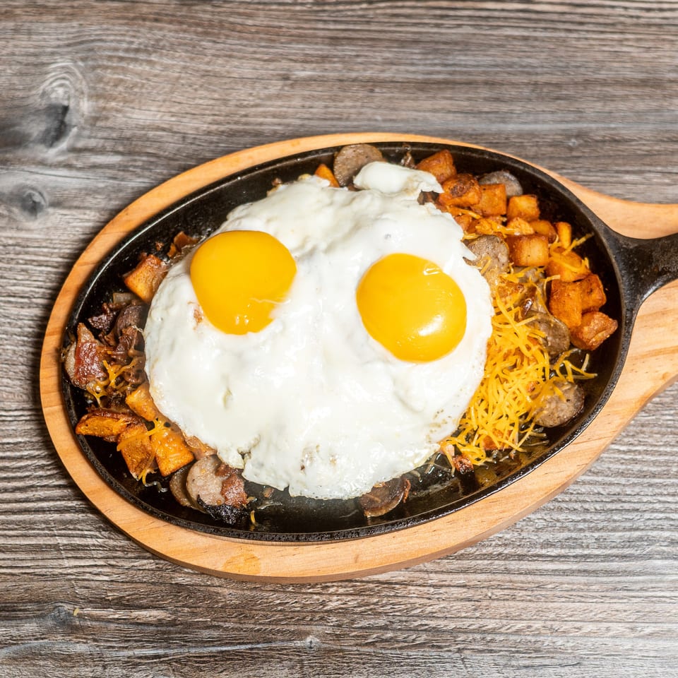 Breakfast skillet with two fried eggs, sausage, potatoes, and shredded cheese on a wooden table.