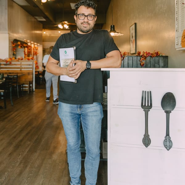 A man in glasses and a black shirt holds menus, leaning on a white counter in a restaurant.