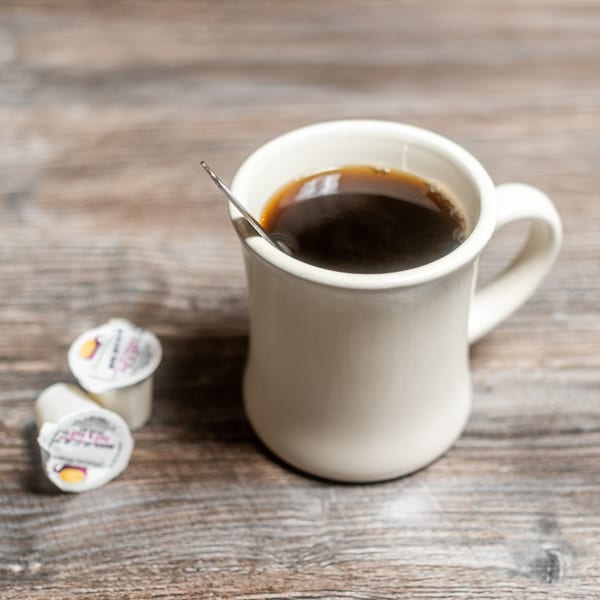 White mug of black coffee with a spoon and two creamers on a wooden table.