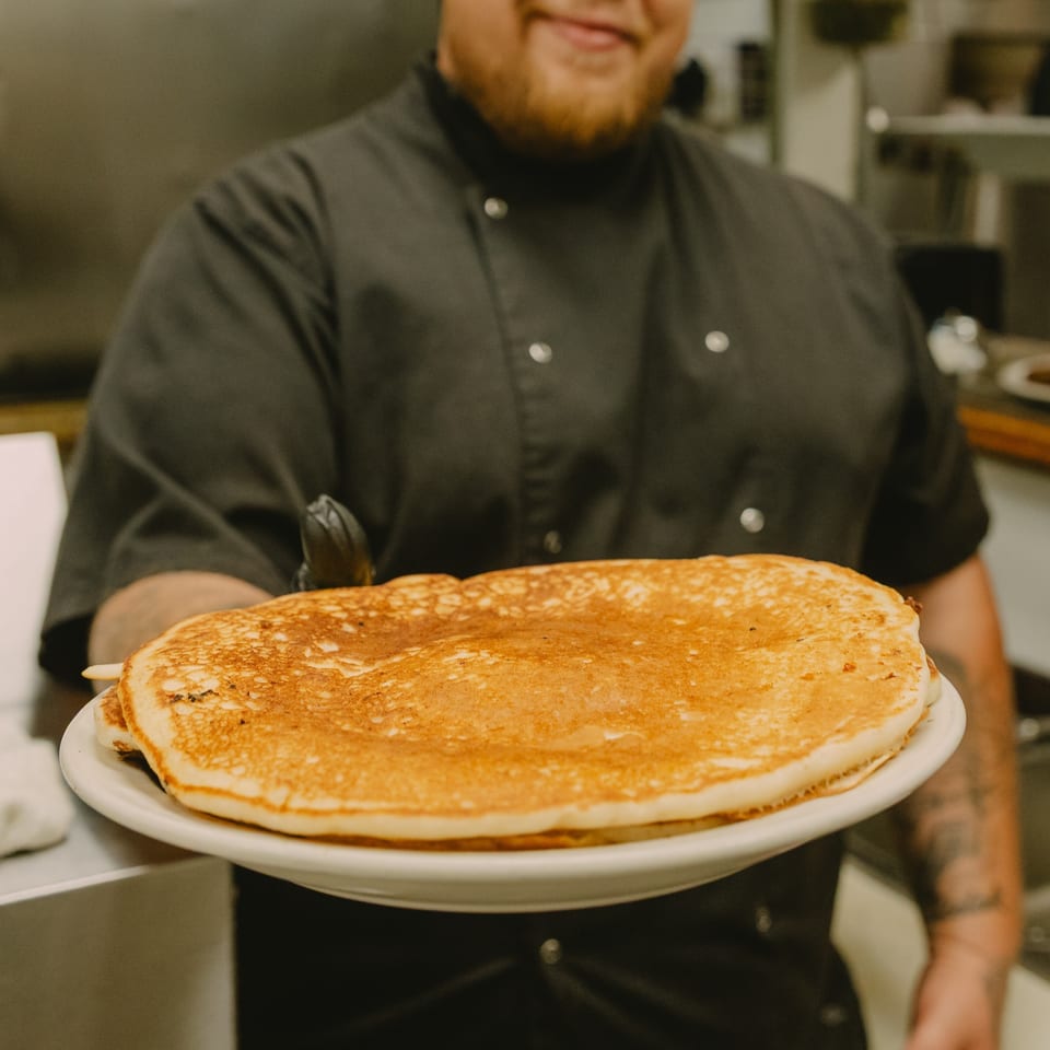 Chef in a black uniform and green cap holding a plate of golden-brown pancakes.