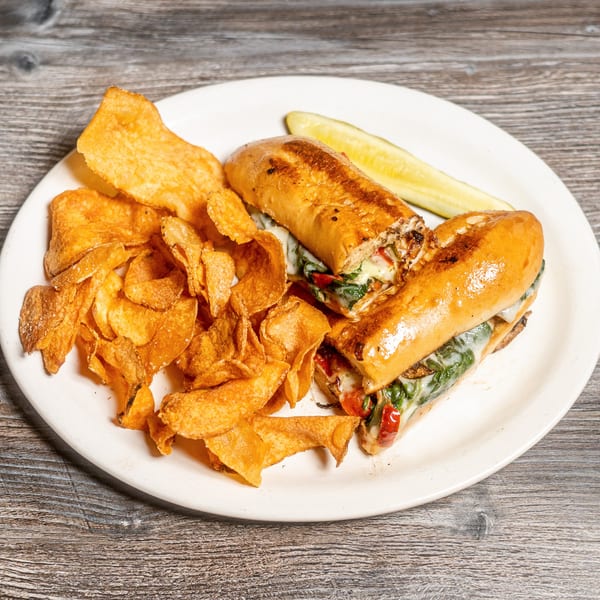 A grilled sub sandwich with melted cheese and vegetables, served with crispy chips and a pickle.