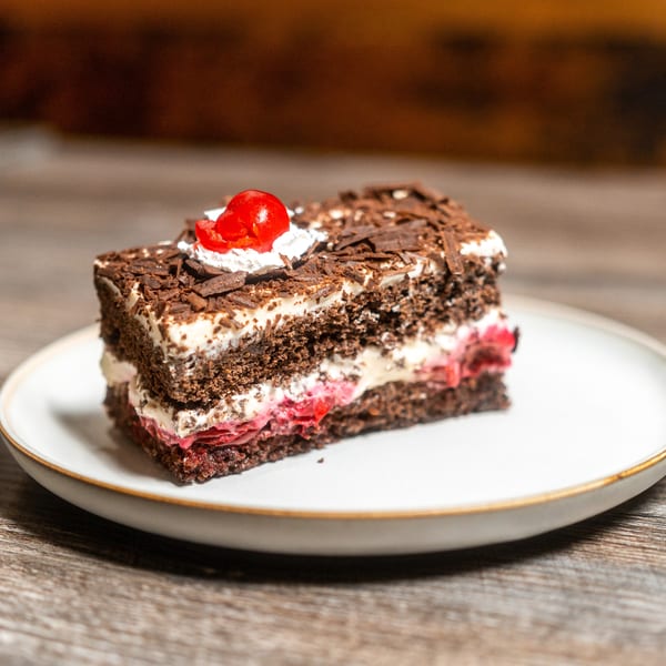 A slice of Black Forest cake with chocolate shavings, whipped cream, and a cherry on a white plate.